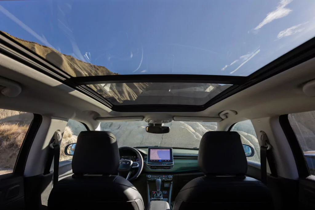Blick von der Rückbank eines Autos mit Panoramadach auf zwei Vordersitze, ein digitales Armaturenbrett und eine felsige Landschaft unter einem strahlend blauen Himmel.