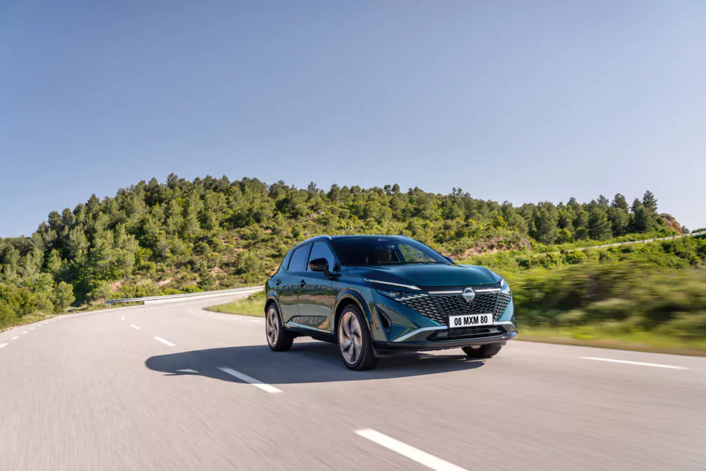 Ein blaugrüner Peugeot 308 fährt auf einer kurvenreichen Straße durch eine malerische, grüne Landschaft mit Hügeln und Bäumen unter einem klaren blauen Himmel.