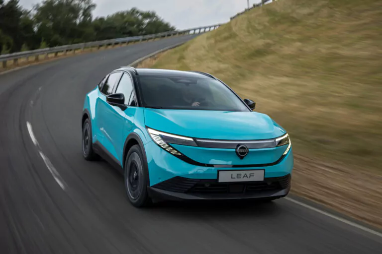 Ein hellblaues Elektroauto mit der Aufschrift LEAF fährt schnell über eine kurvenreiche Straße, die von grasbewachsenen Hängen und Bäumen gesäumt ist, unter einem bewölkten Himmel.