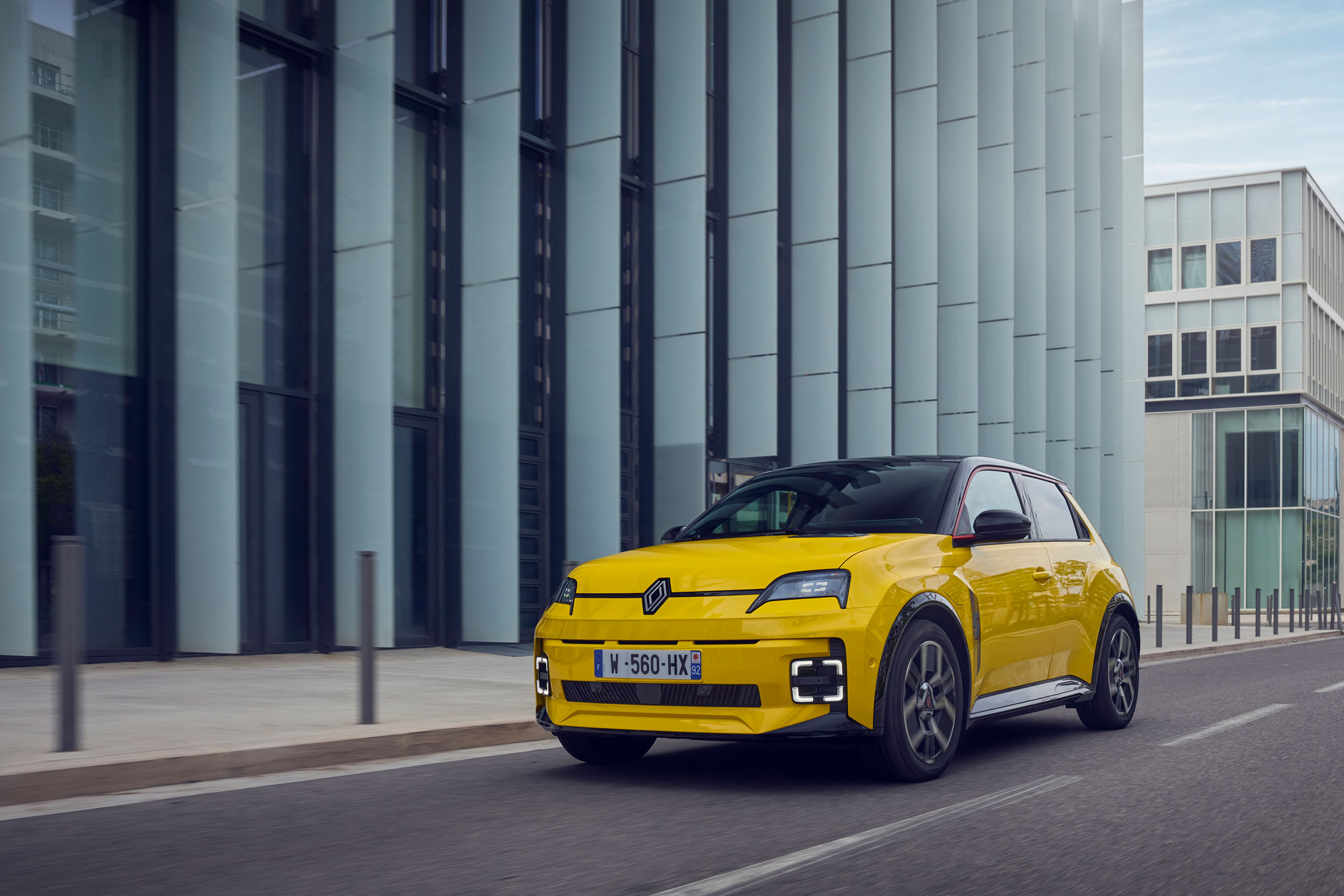 Ein leuchtend gelbes neu-renault-5-e-tech-angebot Elektroauto fährt auf einer Stadtstraße neben einem modernen Glasgebäude unter Tageslicht.
