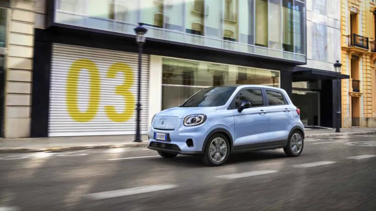 Ein Leapmotor T03 fährt auf einer Stadtstraße vorbei an modernen Gebäuden mit großen Fenstern und einem mit einer gelben 03 bemalten Garagentor.