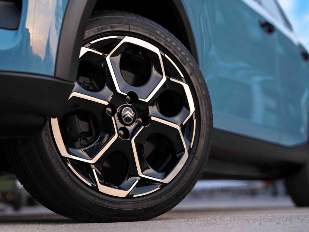 Nahaufnahme eines modernen Autorads mit einem einzigartigen geometrischen Felgendesign in Schwarz und Silber, montiert an einem blauen Citroen C3 You, der auf einer Betonfläche geparkt ist.