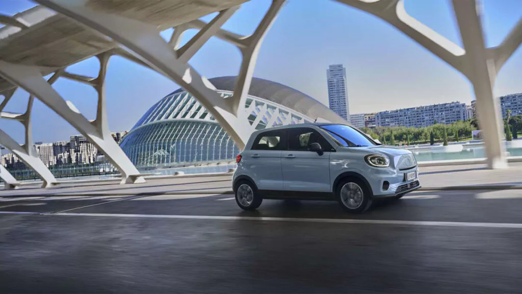 Ein hellblauer Leapmotor T03, fährt auf einer Straße in der Nähe eines modernen Gebäudes, dessen markante weiße architektonische Struktur im Vordergrund zu sehen ist.