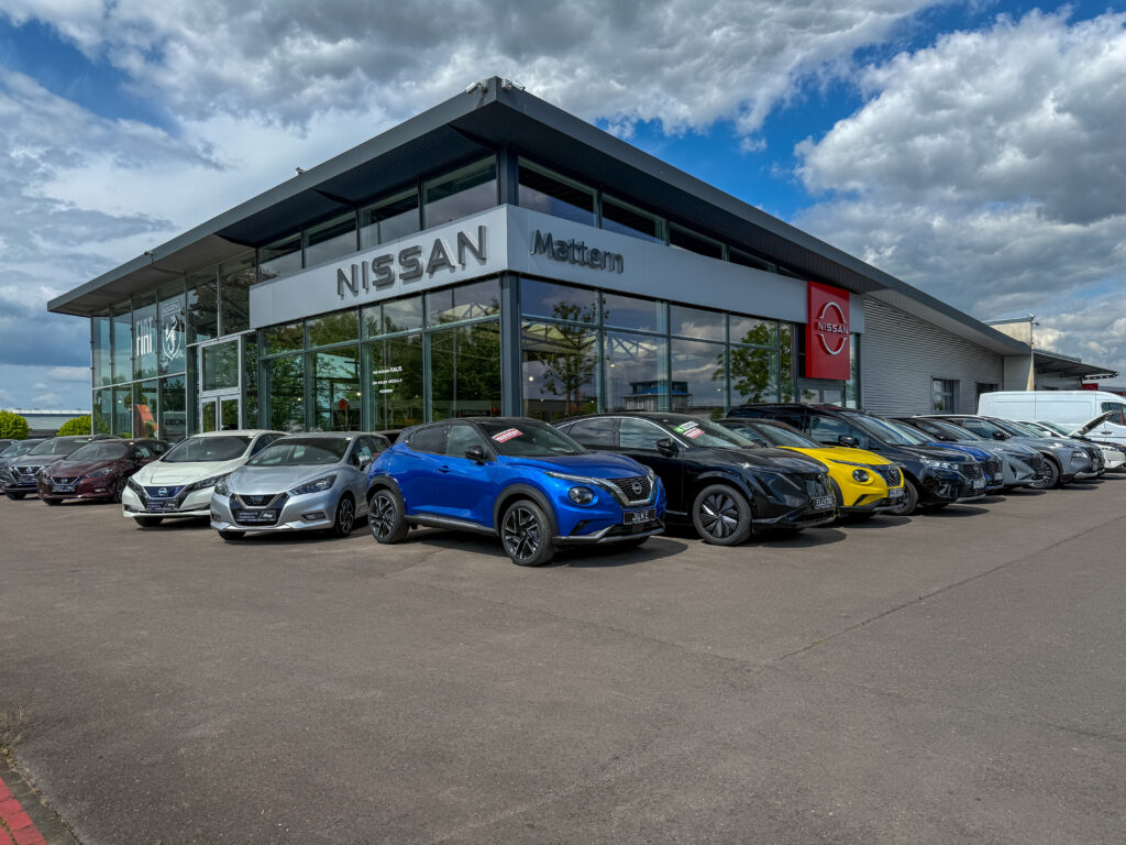 Vor einem modernen Nissan-Autohaus mit großen Glasfenstern steht bei teilweise bewölktem Himmel eine Reihe von Autos, darunter ein leuchtend blauer Wagen.