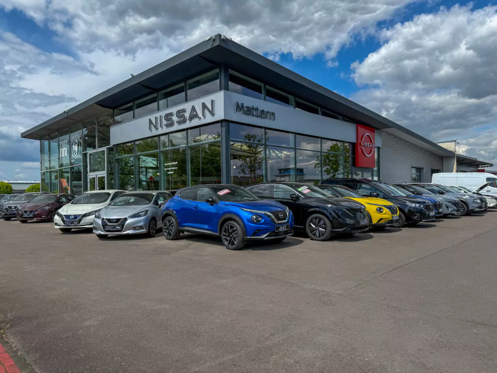 Vor einem modernen Nissan-Autohaus mit großen Glasfenstern steht bei teilweise bewölktem Himmel eine Reihe von Autos, darunter ein leuchtend blauer Wagen.