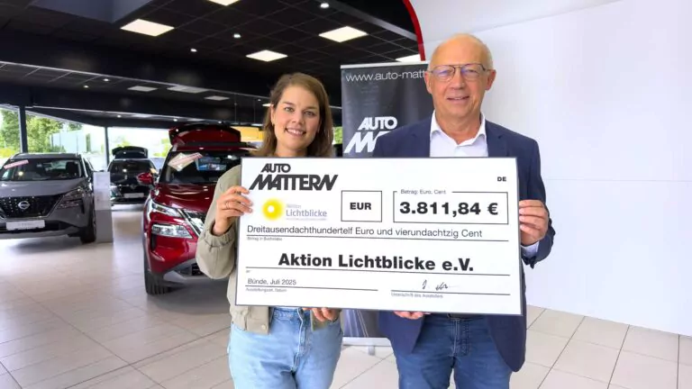 Eine Frau und ein Mann stehen in einem Autohaus und halten einen großen Spendenscheck über 3.811,84 Euro, ausgestellt auf die Aktion Lichtblicke e.V. von Auto Mattern. Im Hintergrund sind Autos und ein Transparent zu sehen.