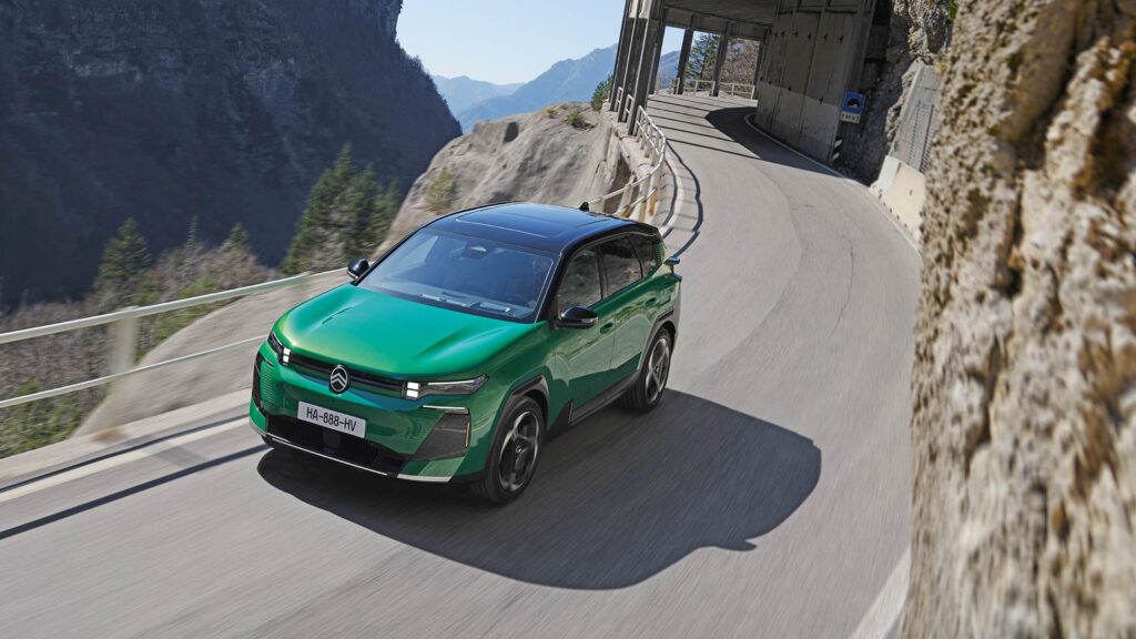 Ein grüner Citroën-Geländewagen fährt während des Aktionswochenendes auf einer kurvenreichen Bergstraße, die von einer Felswand und Metallgeländern begrenzt wird. Im Hintergrund sind bei klarem Himmel Berge und Bäume zu sehen.