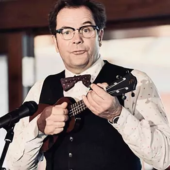 Ein Mann, der eine Brille, eine gepunktete Fliege und eine Weste trägt, spielt eine Ukulele und singt während des Aktionswochenendes in ein Mikrofon.