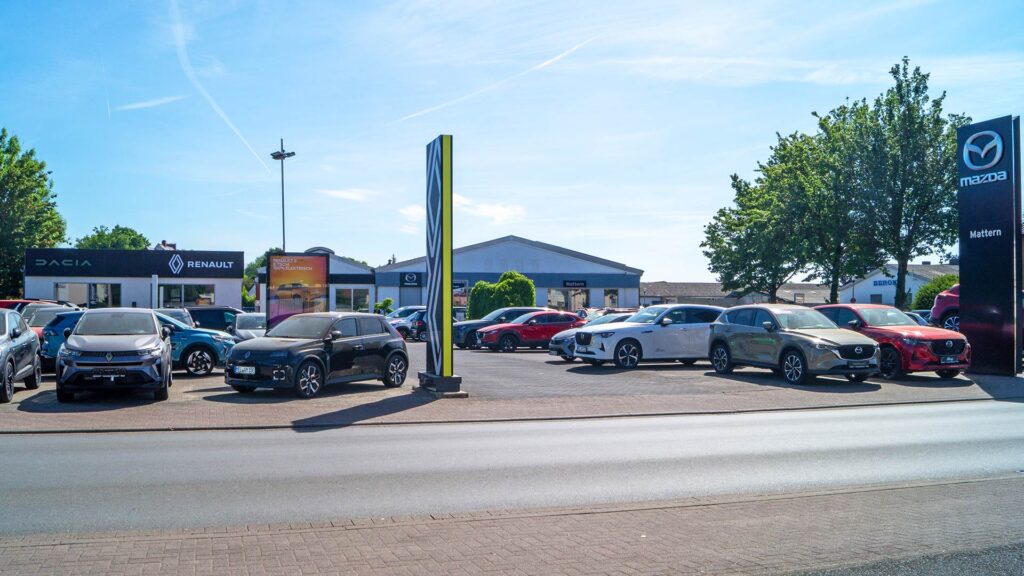 Auf dem Parkplatz eines Autohauses werden unter einem sonnigen blauen Himmel während eines Aktionswochenendes verschiedene Neuwagen ausgestellt. An den Gebäuden im Hintergrund sind Schilder für Mazda, Dacia und Renault zu sehen. Im Vordergrund sind Bäume und eine asphaltierte Straße zu sehen.