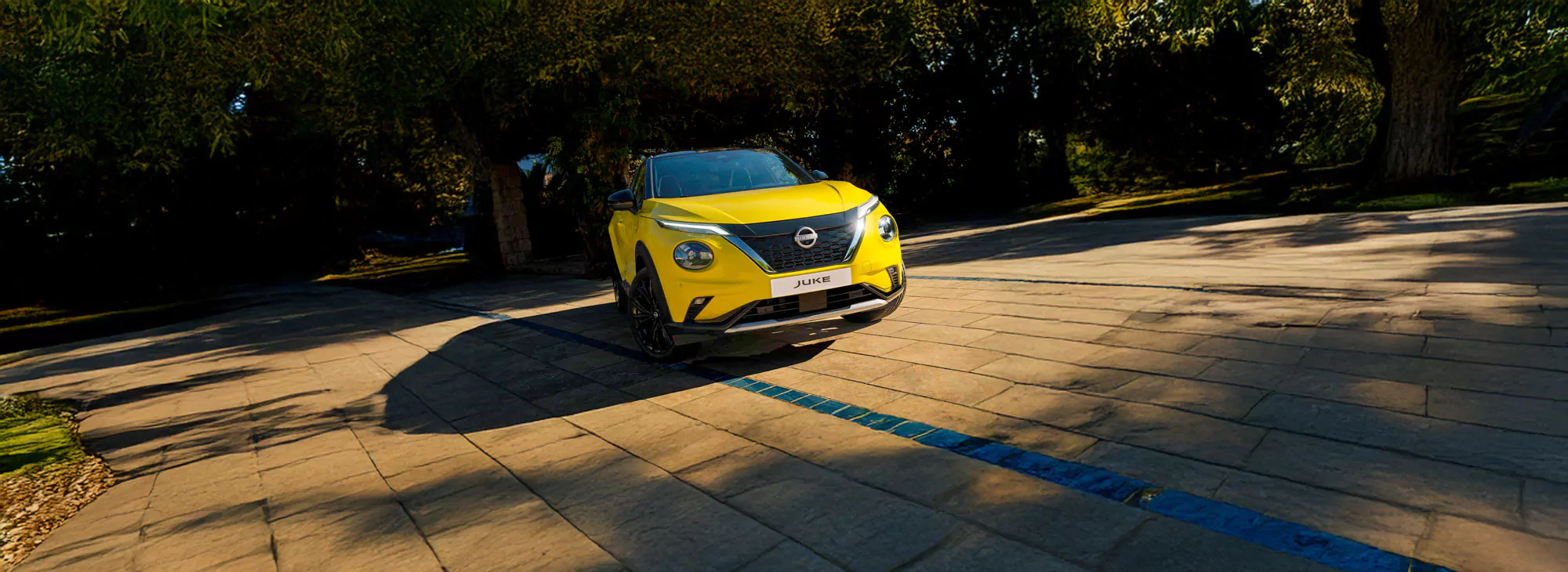 Ein leuchtend gelber Nissan Juke fährt auf einem sonnenbeschienenen, gepflasterten Weg, der von üppigen, belaubten Bäumen umgeben ist und einen Schatten auf den Boden wirft.