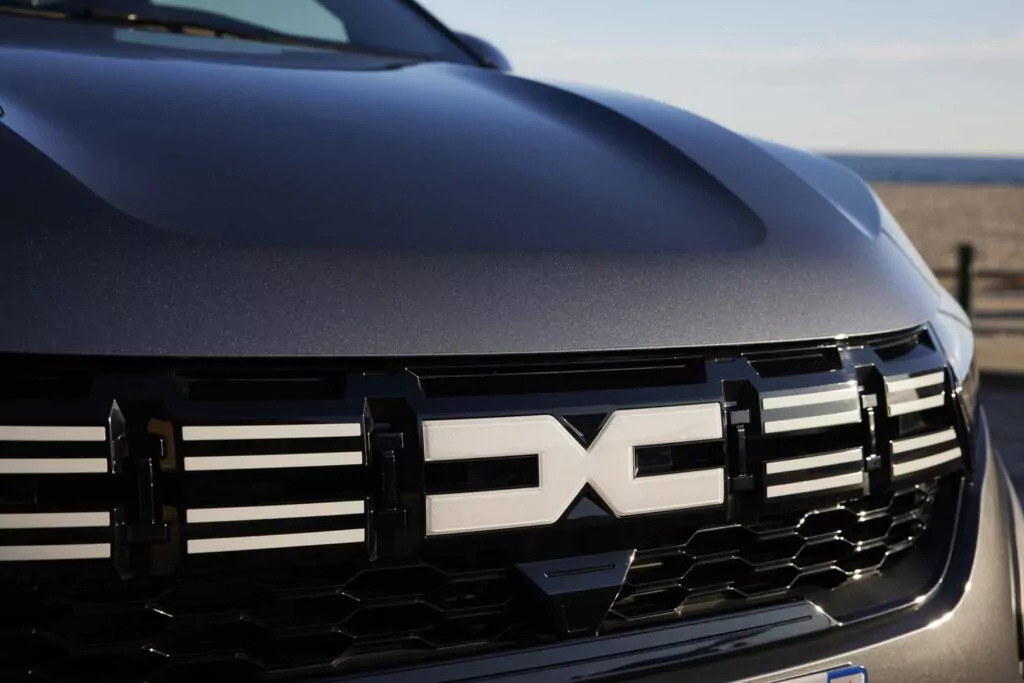 Nahaufnahme des vorderen Kühlergrills eines Autos mit einem auffälligen metallischen Dacia-Logo und geometrischen Designelementen. Die Motorhaube und die Windschutzscheibe sind sichtbar, mit einem unscharfen Strand im Hintergrund - perfekt für die Präsentation des Dacia Jogger Essential Leasing.