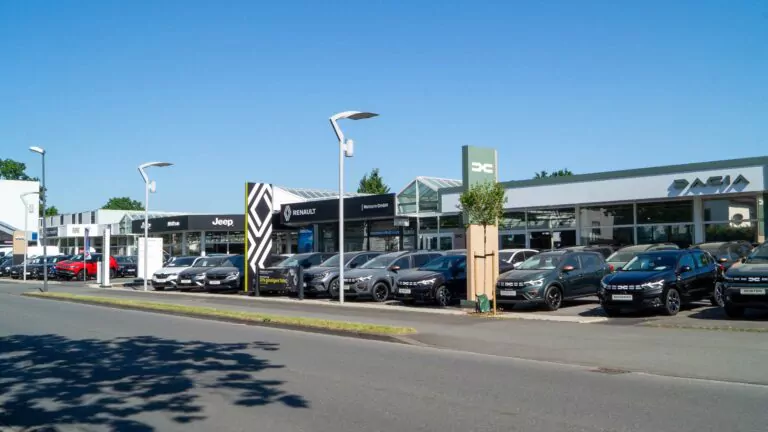 Ein Autohaus mit mehreren Marken, darunter Jeep, Renault und Dacia, zeigt mehrere schwarze und graue Fahrzeuge, die vor einem modernen Gebäude unter einem klaren blauen Himmel geparkt sind.