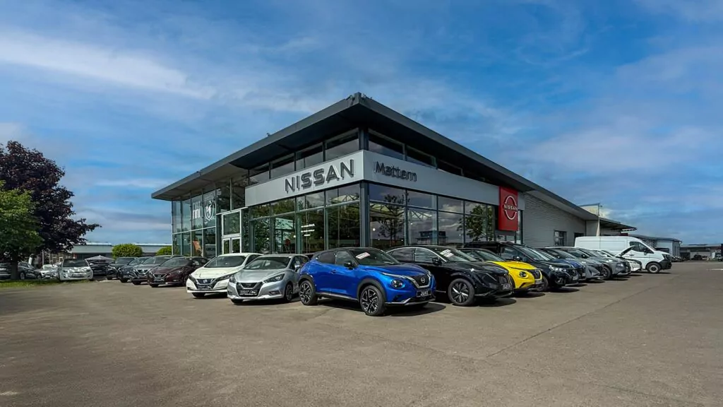 Ein Autohaus mit einer Glasfront, auf der das Nissan-Logo prangt. Mehrere geparkte Autos, darunter ein blaues und ein gelbes Fahrzeug, sind vor dem Gebäude unter einem teilweise bewölkten Himmel aufgereiht.