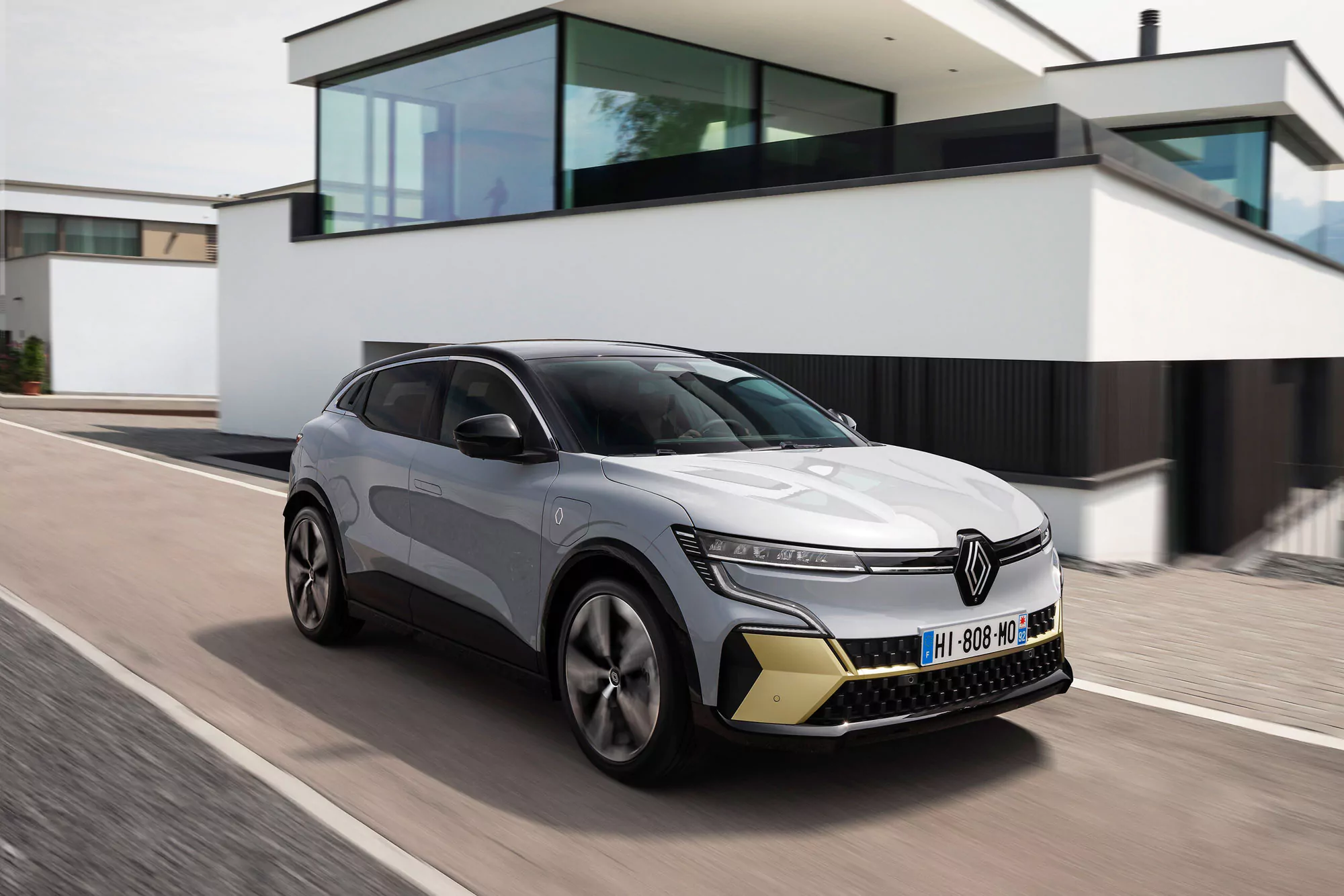 Ein silberner Renault-Elektro-Geländewagen fährt auf einer Vorstadtstraße vor modernen weißen Häusern mit großen Glasfenstern.