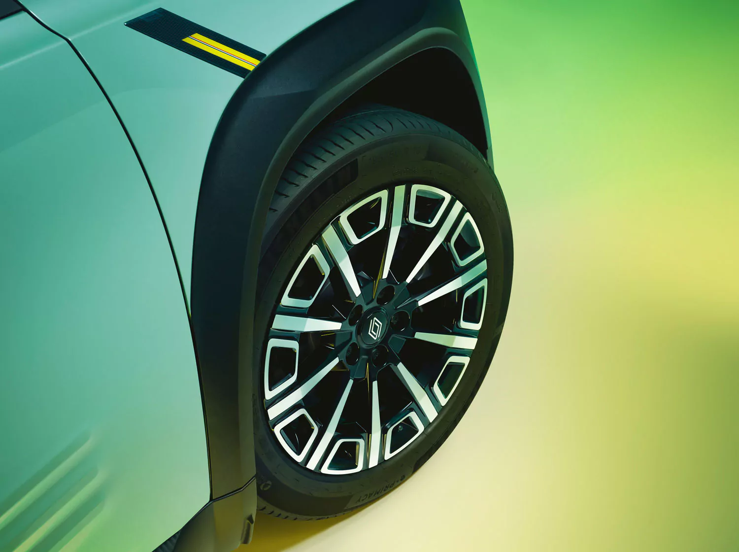 Nahaufnahme des Vorderrads und des Reifens eines Autos mit einer geometrischen schwarz-silbernen Felge und einem grün-gelben Hintergrund mit Farbverlauf. Ein Teil der grün-blauen Karosserie des Fahrzeugs und ein gelber Streifen sind sichtbar.