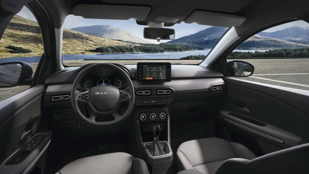 Blick von den Vordersitzen eines Dacia-Fahrzeugs auf das Armaturenbrett, das Lenkrad, die Mittelkonsole und den Touchscreen - perfekt für diejenigen, die Dacia Jogger Essential Leasing in Betracht ziehen - mit einem malerischen See und Bergen durch die Windschutzscheibe.