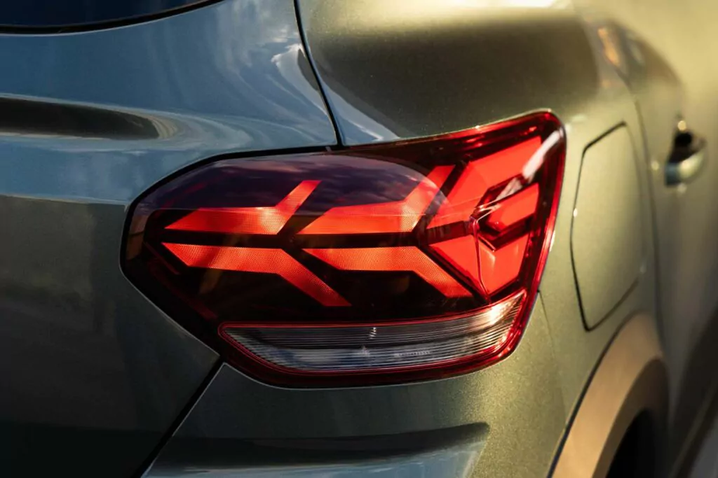 Nahaufnahme des linken hinteren Rücklichts eines modernen Autos mit einem geometrischen, beleuchteten roten Muster. Der Dacia Sandero Essential für 89 € mtl. leasen erstrahlt in Grünmetallic, mit glatten, geschwungenen Karosserielinien und Sonnenlicht, das sich auf seiner Oberfläche spiegelt.