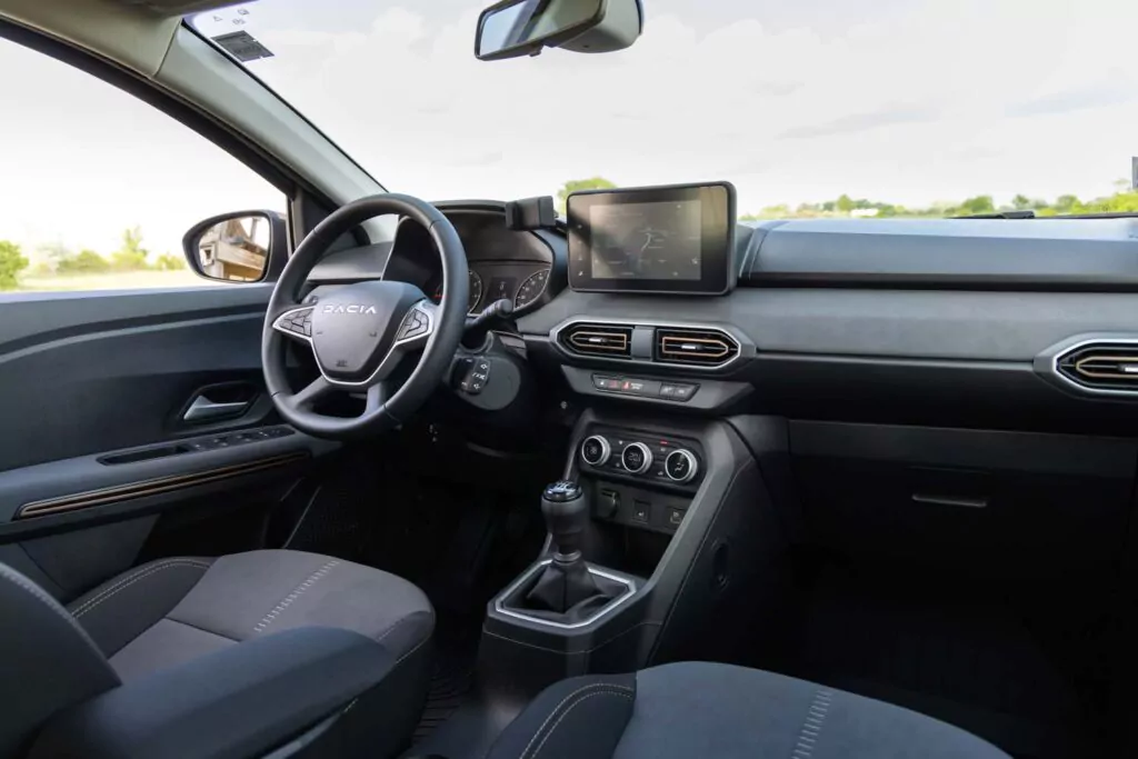 Ein modernes Interieur mit Dacia-Lenkrad, Touchscreen-Display, manuellem Schalthebel und schwarzen Sitzen mit dezenten Details - das erleben Sie im Dacia Sandero Essential für 89 € mtl. Leasing, mit Blick ins Grüne durch die Fenster.