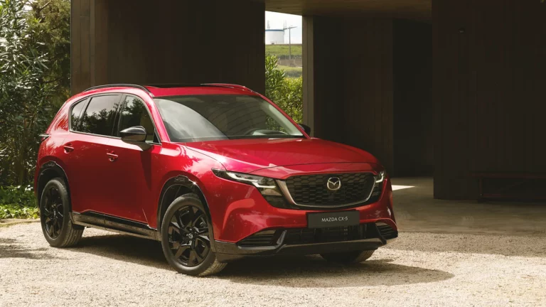 Ein roter Mazda CX-5 SUV ist auf einer Kiesauffahrt in der Nähe eines modernen Gebäudes geparkt, im Hintergrund sind Bäume und Grünflächen zu sehen.