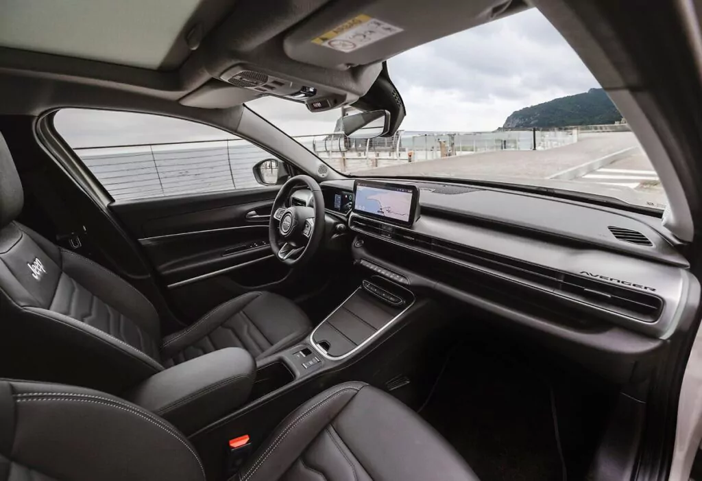 Ansicht eines modernen Autoinnenraums mit schwarzen Ledersitzen, einem digitalen Armaturenbrett, einem Lenkrad und einem zentralen Display mit Navigationsfunktion. Die Szene draußen zeigt einen bewölkten Himmel und Berge im Hintergrund.