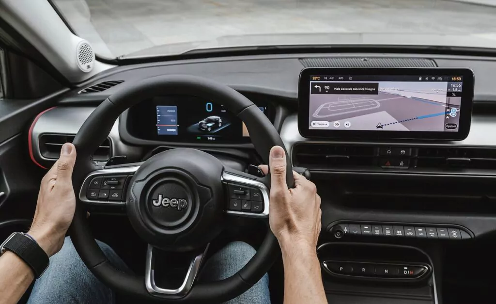 Eine Person hält das Lenkrad eines Jeeps mit beiden Händen fest. Das Armaturenbrett zeigt digitale Anzeigen und eine Navigationskarte auf einem großen Touchscreen an. Der Innenraum ist modern und schnittig.
