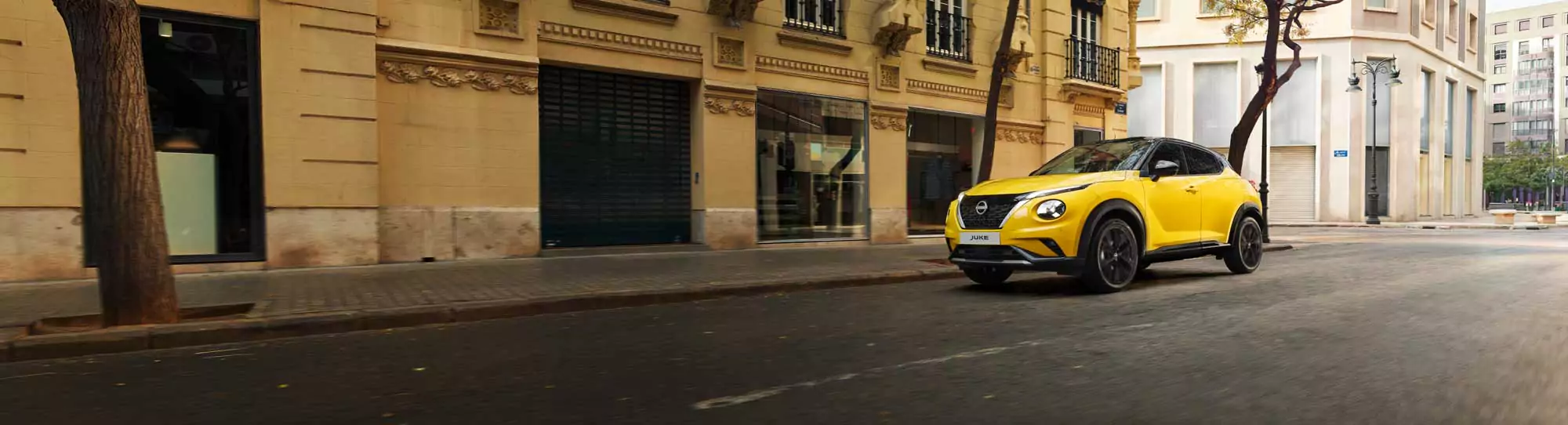 Ein leuchtend gelber Nissan Juke Angebot fährt eine ruhige Stadtstraße entlang, die von beigefarbenen historischen Gebäuden und Bäumen gesäumt ist. Die Bürgersteige sind leer und das sanfte Sonnenlicht beleuchtet die Szene.