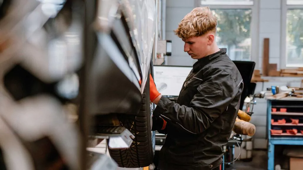 Ein Mechaniker in schwarzer Uniform und roten Handschuhen inspiziert oder repariert ein Fahrzeug, das auf einer Hebebühne in einer Werkstatt mit Werkzeugen und Regalen im Hintergrund steht.