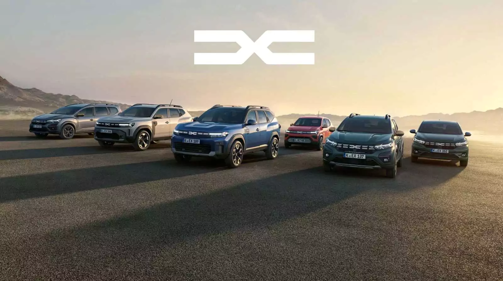 Sechs Dacia-Geländewagen sind in einer Reihe in einer flachen, offenen Landschaft bei Sonnenuntergang geparkt, mit einem großen weißen Dacia-Logo über ihnen im Himmel. Im Hintergrund sind Berge und ein klarer Himmel zu sehen.