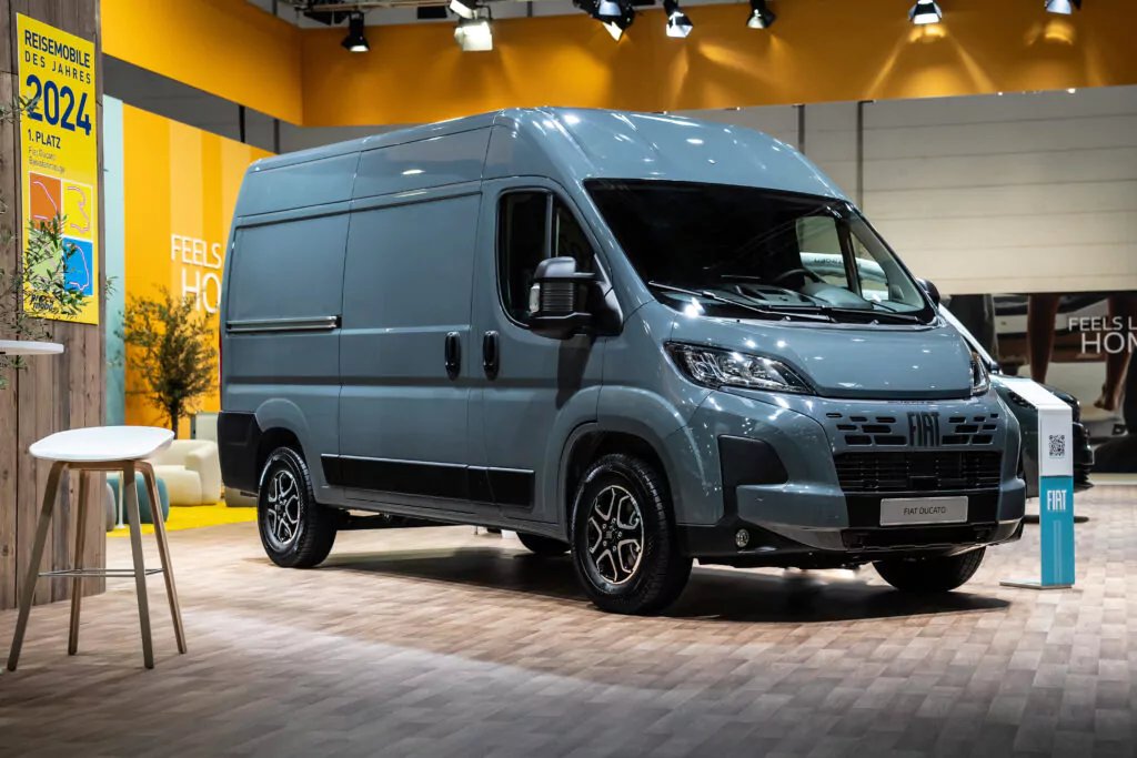 Ein grauer Fiat E-Ducato Elektro-Van steht in einem Innenraum auf einem Holzboden, mit einer gelben Wand und Informationsschildern im Hintergrund.