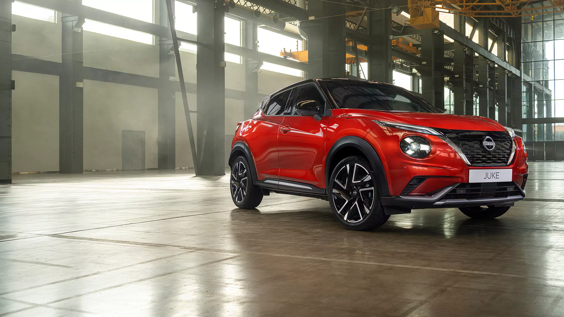Ein roter Nissan Juke SUV steht in einer geräumigen Lagerhalle im Industrial-Stil mit großen Fenstern und Metallträgern und bringt den modernen Charme des aktuellen Nissan Juke-Angebots perfekt zur Geltung.