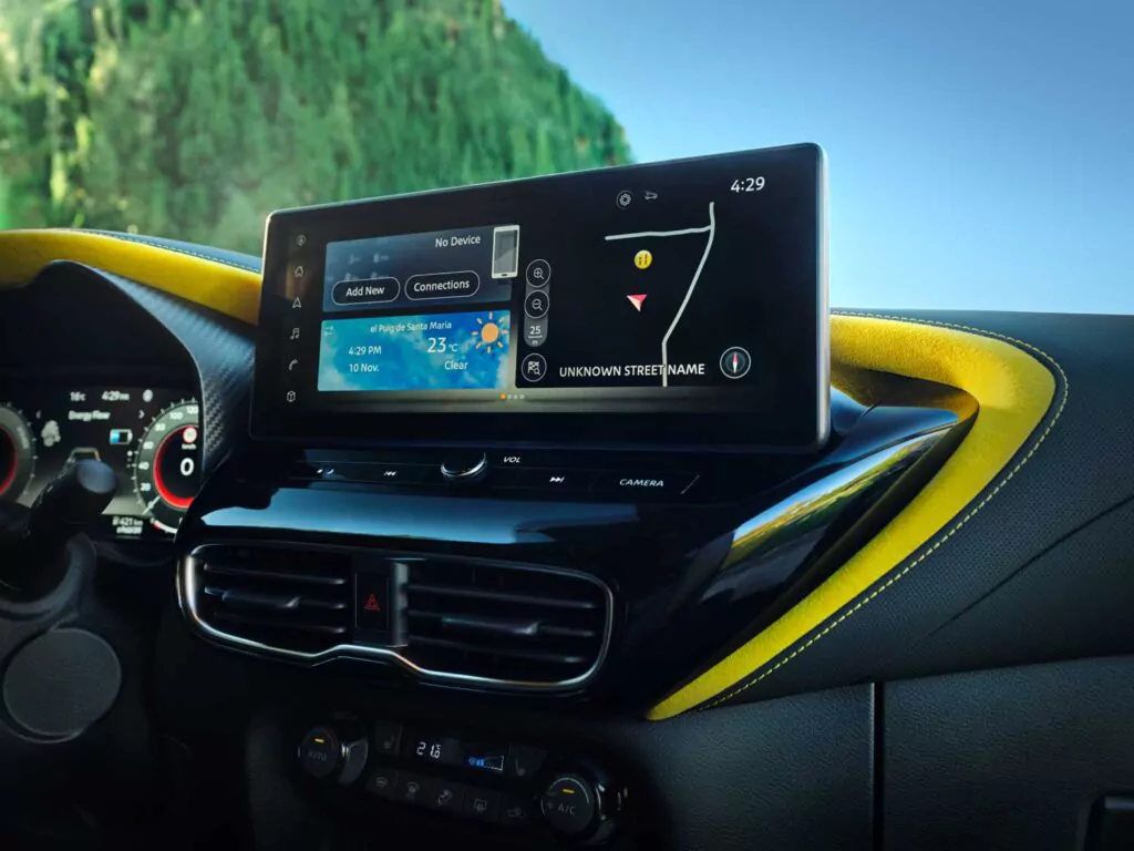 Nahaufnahme des Armaturenbretts eines Nissan Juke Angebot mit einem digitalen Display, das Navigation, Mediensteuerung und Klimainformationen anzeigt. Der Innenraum ist in Schwarz und Gelb gehalten, und durch das Fenster ist ein Wald zu sehen.