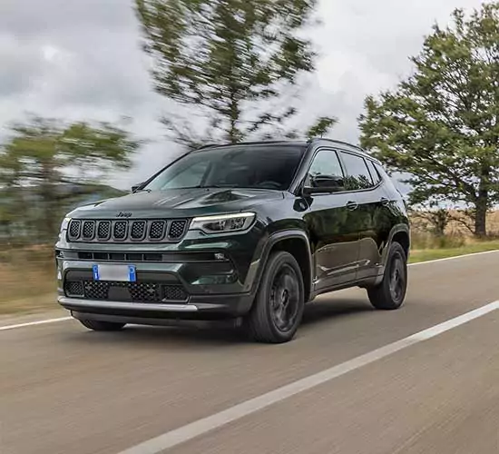 Ein schwarzer Jeep-Geländewagen fährt auf einer Landstraße, umgeben von Bäumen und einem bewölkten Himmel im Hintergrund.