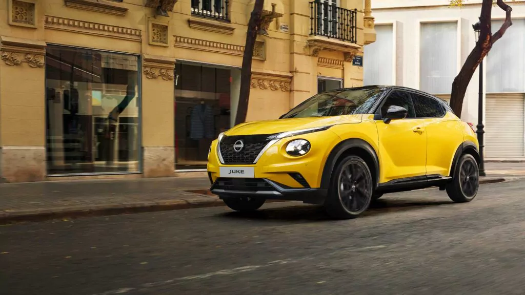 Ein gelber Nissan Juke, ein kompakter Geländewagen, fährt eine Straße entlang, die von hellbraunen Häusern mit Balkonen und verzierten Details gesäumt ist, und präsentiert das neueste Nissan Juke Angebot in einer städtischen Umgebung.