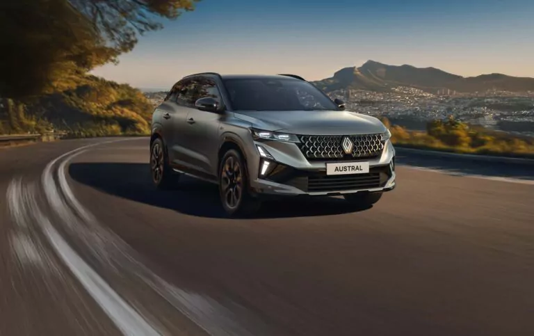 Ein silberner Renault Austral SUV, der bei Renault Austral Leasing erhältlich ist, fährt auf einer kurvenreichen Straße, umgeben von grünen Hügeln und einer entfernten Stadt unter einem klaren Sonnenuntergangshimmel.