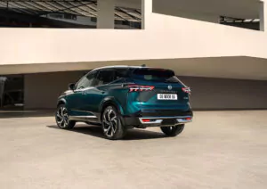 Ein tealfarbener Nissan Qashqai N-Connecta SUV ist auf einer glatten, sonnenbeschienenen Betonfläche vor einem modernen weißen Gebäude mit geschwungenen architektonischen Elementen geparkt. Das Fahrzeug wird von der hinteren linken Seite aus betrachtet.