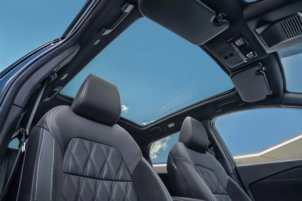 Blick aus dem Innenraum eines Nissan Qashqai N-Connecta mit schwarzen Ledersitzen mit weißen Nähten und einem großen Panorama-Schiebedach, das einen klaren blauen Himmel mit ein paar Wolken zeigt.