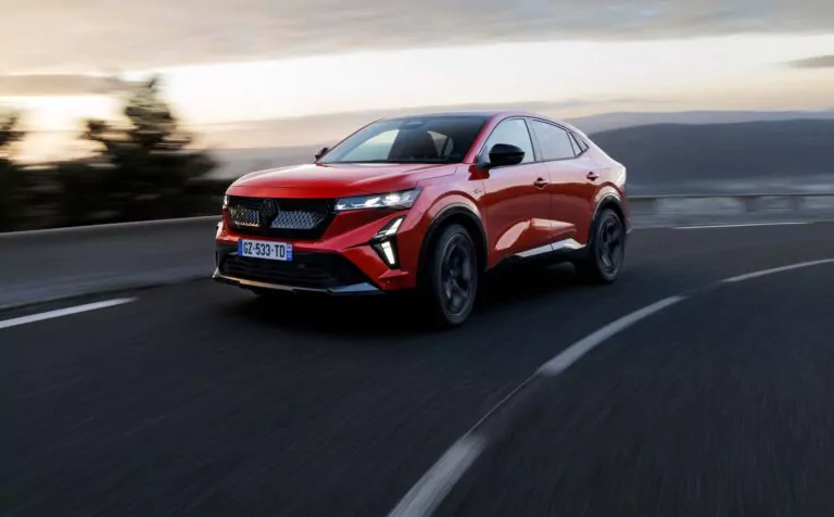 Ein roter Renault Rafale Vollhybrid-Geländewagen mit schnittigem, modernem Design fährt bei Sonnenuntergang auf einer kurvenreichen Straße, mit verschwommenen Bäumen und Hügeln im Hintergrund, die ein Gefühl von Geschwindigkeit vermitteln - perfekt für alle, die über ein Leasing nachdenken.