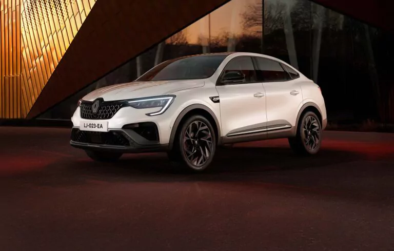 Ein weißer Renault Arkana SUV parkt auf einem dunklen Bürgersteig vor einem modernen Gebäude mit goldenen, kantigen Paneelen und Glasfenstern, die von einer warmen Beleuchtung angestrahlt werden.