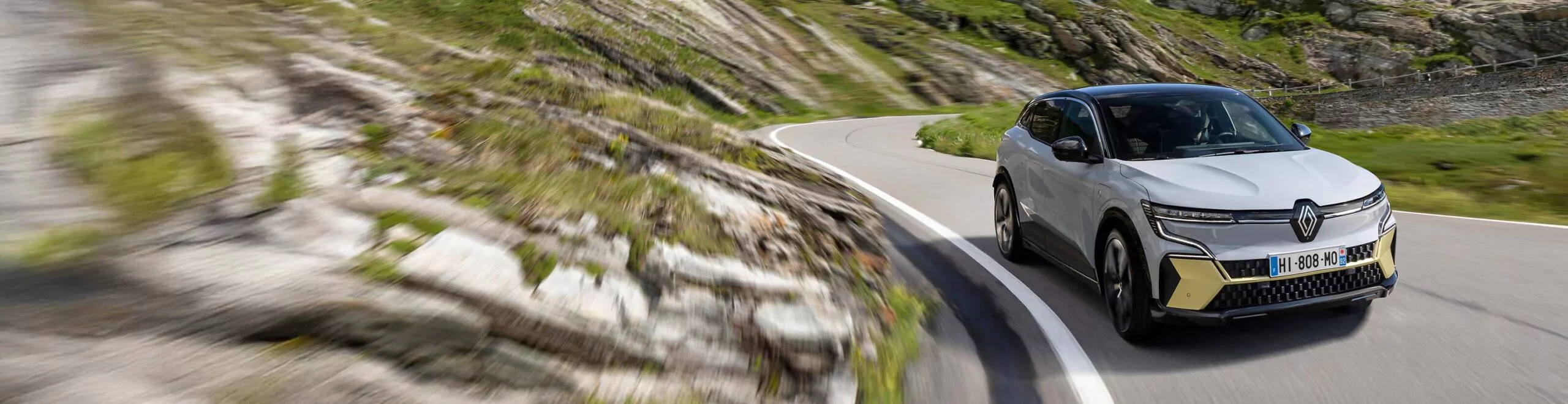 Ein weiß-schwarzer Renault Megane E-Tech mit französischem Kennzeichen fährt auf einer kurvenreichen Bergstraße, umgeben von felsigem Gelände und grünem Gras unter einem strahlenden Himmel - eine ideale Szene für Renault Megane E-Tech Leasing-Fans.