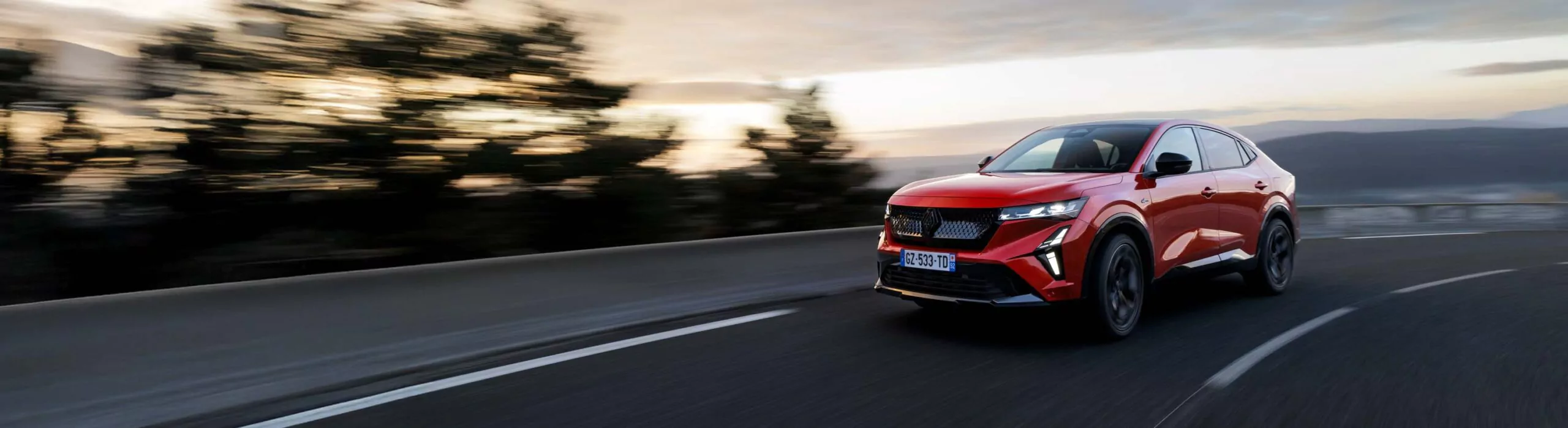 Ein roter Renault Rafale Vollhybrid-Geländewagen fährt bei Sonnenuntergang zügig eine kurvige Straße entlang, mit verschwommenen Bäumen und Himmel im Hintergrund, die Bewegung und Geschwindigkeit suggerieren - perfekt für alle, die Full Hybrid Leasing in Betracht ziehen.