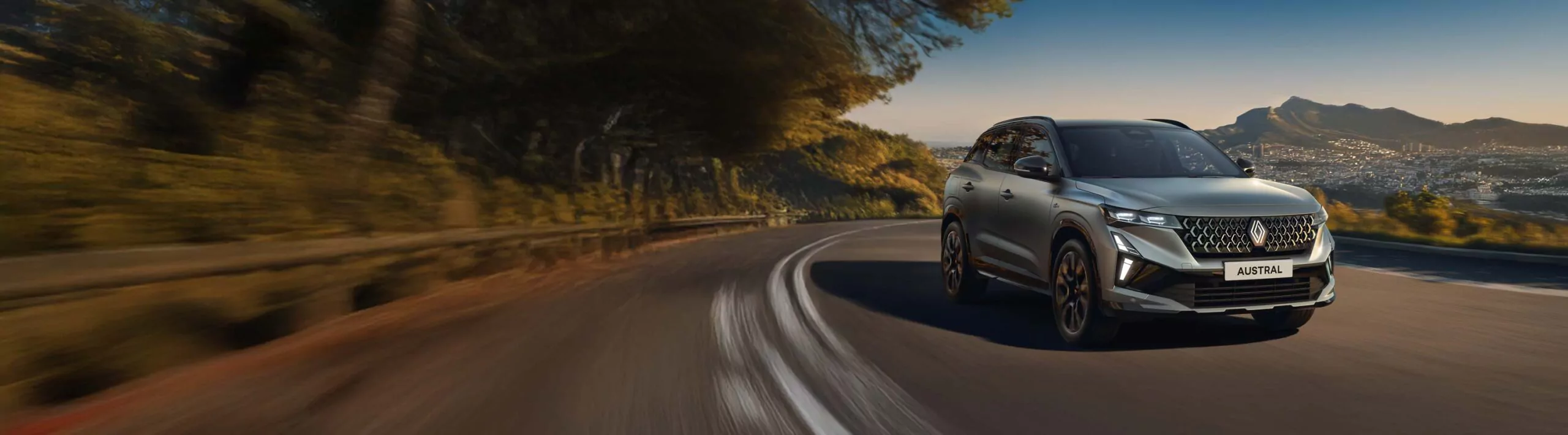 Ein silberner Geländewagen mit der Aufschrift Austral fährt auf einer kurvenreichen Bergstraße, umgeben von Bäumen, mit einer Stadt und Bergen in der Ferne unter einem klaren Himmel - ein Zeichen für die Freiheit von Renault Austral Leasing.