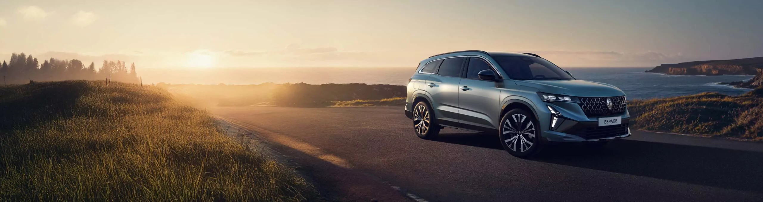 Ein silberner Geländewagen parkt bei Sonnenuntergang auf einer leeren Straße, im Hintergrund sind grasbewachsene Hügel und die Küste bei klarem Himmel zu sehen.