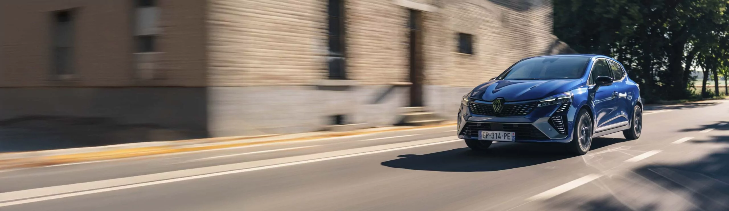 Ein blauer Renault Clio, der über Leasing erhältlich ist, fährt schnell auf einer sonnigen Straße an einem Backsteingebäude vorbei. Die unscharfe Umgebung suggeriert Bewegung, während im Hintergrund Bäume die Straße säumen.