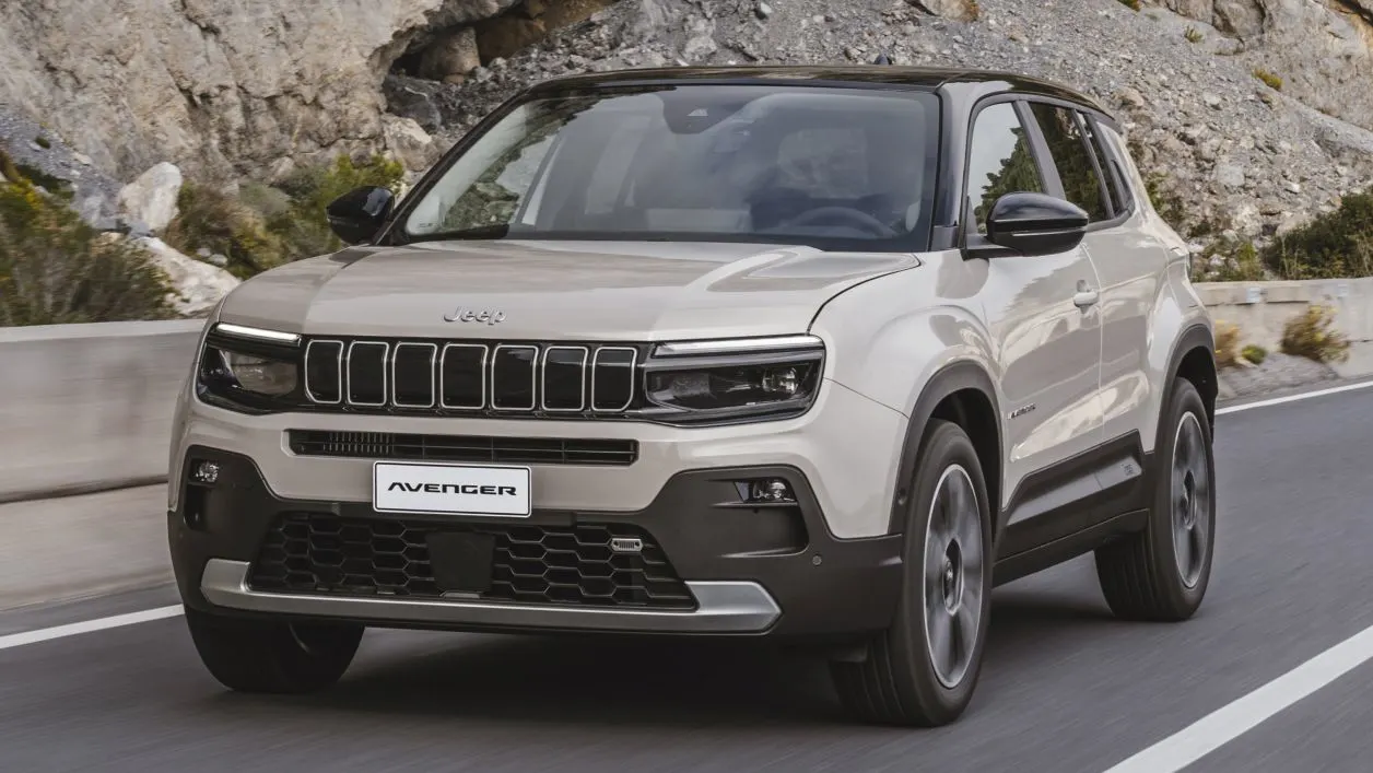Ein beiger Jeep Avenger SUV fährt auf einer asphaltierten Bergstraße, umgeben von felsigem Gelände und Grünflächen. Das Auto hat den typischen Jeep-Kühlergrill und moderne LED-Scheinwerfer.