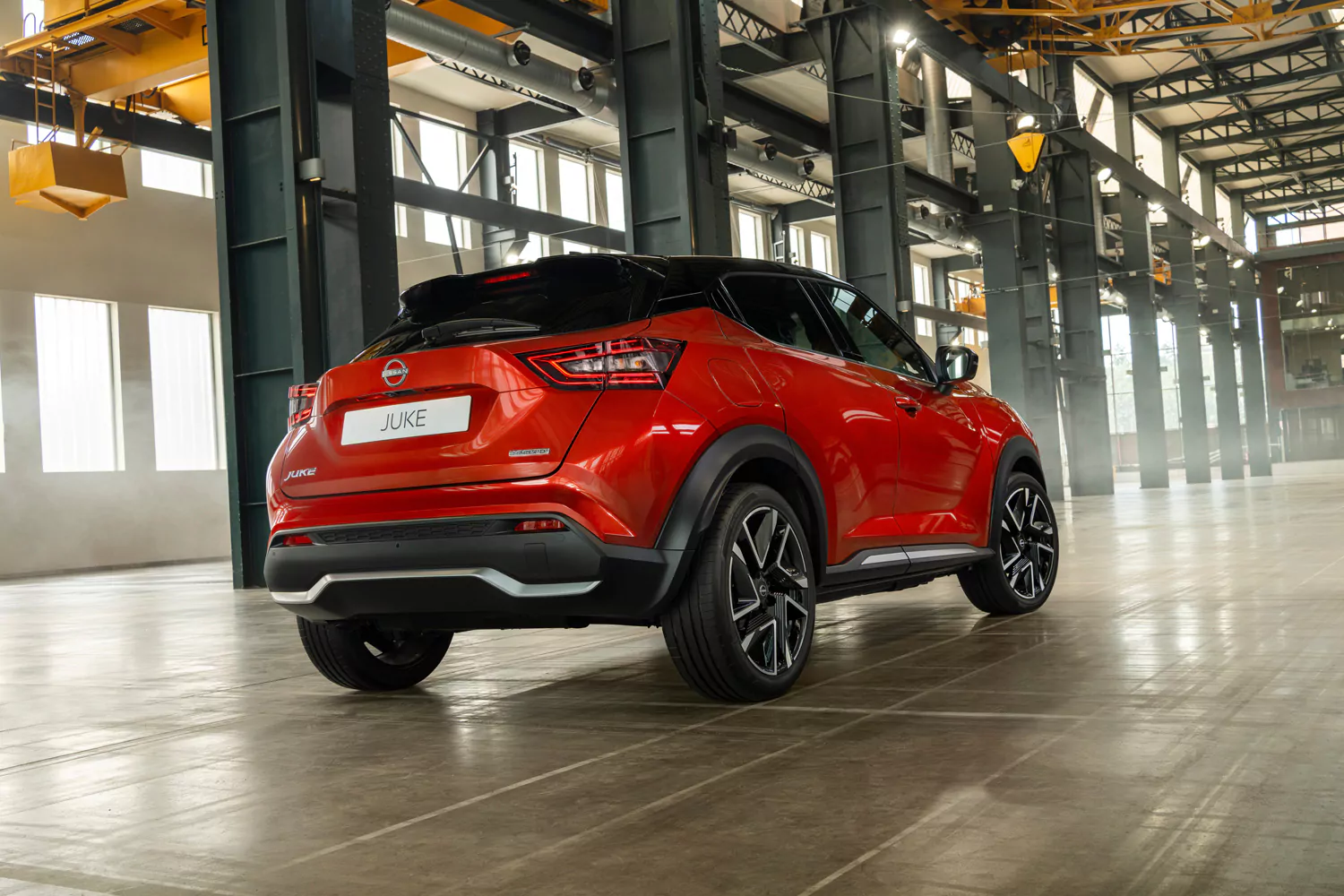 Ein roter Nissan Juke steht in einer großen Halle im Industriestil mit hohen Decken und Metallträgern. Das Fahrzeug wird von links hinten gezeigt, wodurch sein markantes Design besonders gut zur Geltung kommt – ein echter Blickfang für alle, die sich für das aktuelle Nissan Juke N-Connecta-Angebot interessieren.