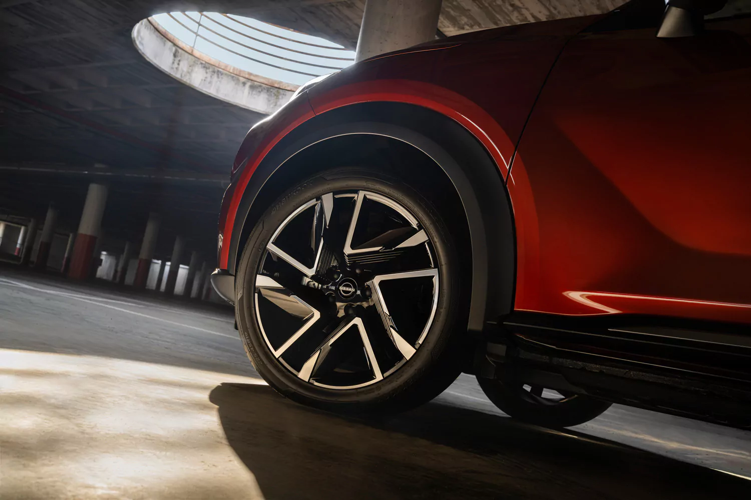 Nahaufnahme des Vorderrades eines roten Autos, das in einer schwach beleuchteten Garage geparkt ist, durch deren runde Öffnung in der Decke Sonnenlicht hereinströmt – ein Bild, das die beeindruckende Präsenz des Nissan Juke N-Connecta Angebot einfängt.
