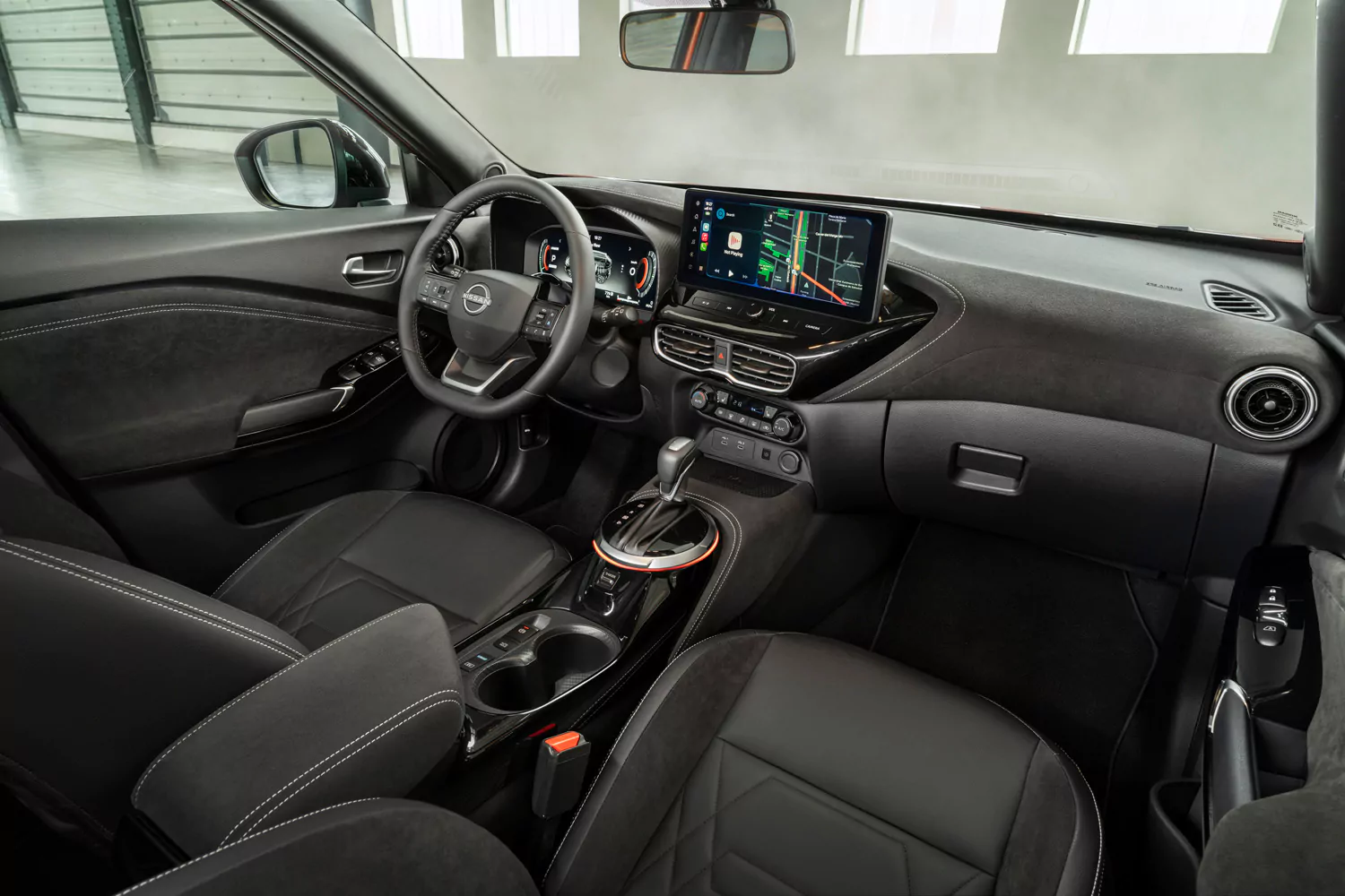 Moderner Fahrzeuginnenraum mit schwarzen Sitzen, einem Touchscreen-Display mit Navigationsanzeige und einem Lenkrad. Das Armaturenbrett verfügt über digitale Bedienelemente; zwischen den Vordersitzen sind der Schalthebel und die Getränkehalter zu sehen. Große Fenster lassen Tageslicht herein – ein perfektes Beispiel für den Stil und die Innovation des Nissan Juke N-Connecta.