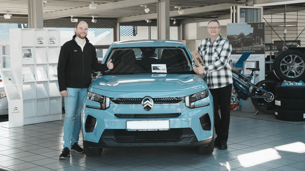 Zwei Männer stehen lächelnd neben einem hellblauen Citroën-Auto in einem hellen Ausstellungsraum. Beide zeigen den Daumen nach oben, und im Hintergrund sind Autoauslagen und Reifen zu sehen.