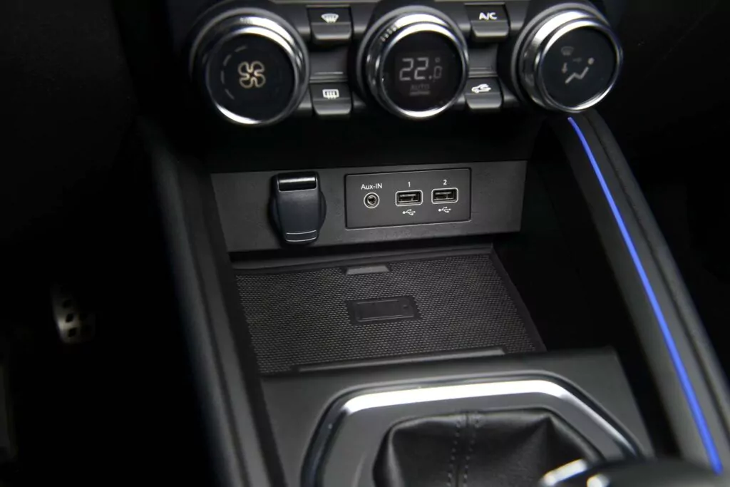Die Nahaufnahme der Mittelkonsole eines Renault Clio Leasing zeigt die Drehregler für die Klimatisierung über einem AUX-Eingang, zwei USB-Anschlüssen und einer 12-Volt-Steckdose, neben dem Schalthebel und einer blau beleuchteten Akzentlinie.