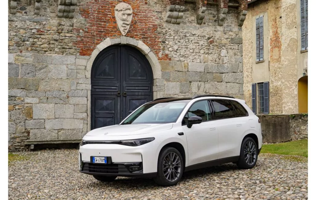 Ein weißer Geländewagen parkt auf Kopfsteinpflaster vor einem rustikalen Steingebäude mit einer gewölbten schwarzen Tür und einer Ziermaske darüber. Das Gebäude hat Ziegel- und Steinmauern und verschlossene Fenster.